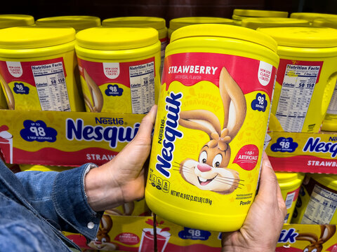Shopper's hand holding a plastic container of Nestl&eacute; Brand Nesquik strawberry  powdered flavoring mix in a supermarket aisle