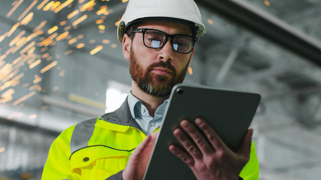 Mature bearded man on construction site. Engineer in uniform and glasses holds tablet background of workflow. An employee notes something in his gadget in workshop. Diversity of proffesions.