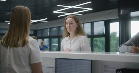 Adult woman stands near reception desk in clinic lobby area and makes appointment with doctor. Female administrator talks with patient at information counter. Medical staff work in modern hospital.