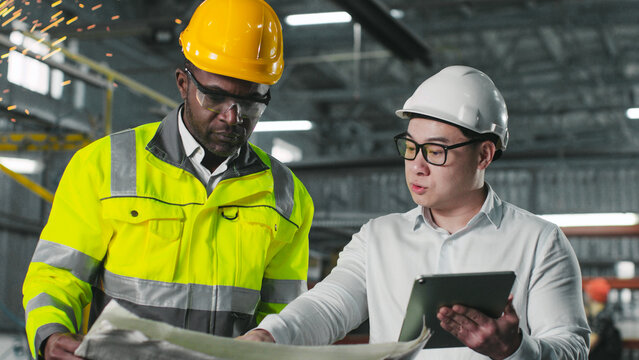 Multiethnic engineers in hads hats and protective clothes walking at construction site. African american engineer holds blueprint. Asian manager use tablet for work. Ethnic diversity