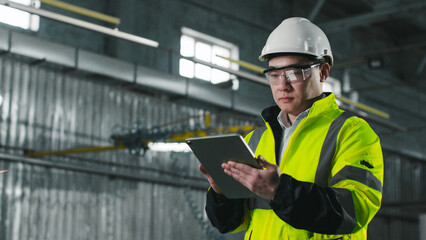 Asian engineer looks at screen of tablet. Worker wearing yellow vest, glasses and hard hat. Male stands on background of workflow. Man at workplace.