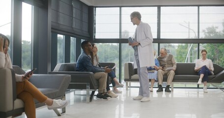 Diverse people sit on sofas in clinic lobby area, wait for appointment with doctor. Doctor speaks with multiethnic couple about medical test results. Waiting area in modern medical center. Healthcare.