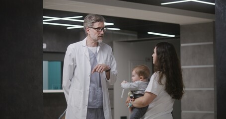 Fototapeta premium Mature doctor discusses medical examination or test results with female patient with little boy. Mother with child and doctor in clinic or medical center lobby after appointment. Healthcare system.