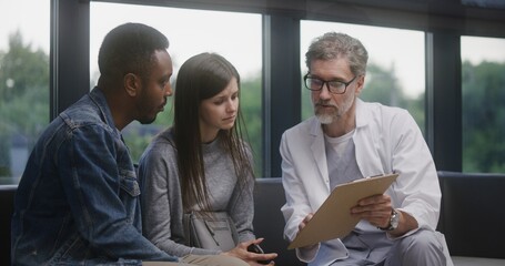 Multiethnic couple sits on sofa in clinic lobby area after appointment with doctor. Doctor talks,...