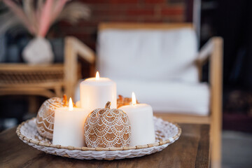Autumn fall cozy mood composition for hygge home decor. Orange pumpkins decorated with mandalas, white burning candles on wicker tray on the coffee table in the loft living room. Selective focus