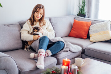 Woman in plaid with tea cup watching movie, TV, petting cat on sofa at home with christmas decoration atmosphere. Lady wear jumper and warm socks. Cozy and comfortable winter concept. Selective focus.