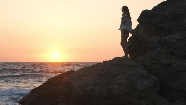 Lonely beautiful woman enjoying sunrise or sunset on the beach at sea on a windy day. Woman on rock in sea