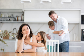 Frustrated wife sitting at kitchen table during family quarrel with angry husband standing behind her. Children are worried about parents quarrel. Son hugs dad, daughter gently leaned against sad mom