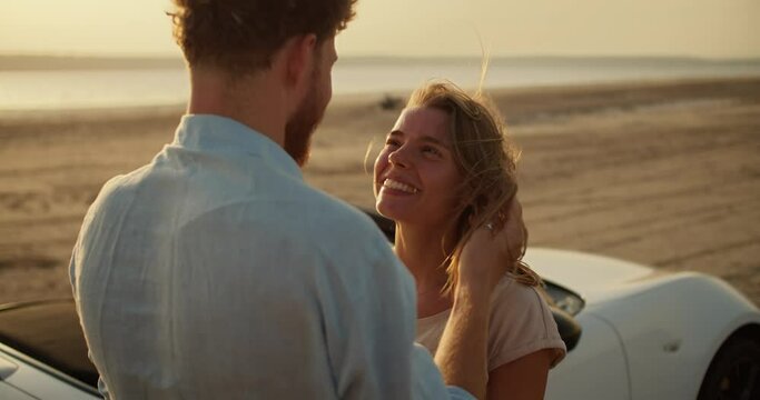 A happy girl is hugging her boyfriend in a blue shirt near a white car. A romantic summer meeting against the river and the yellow sky