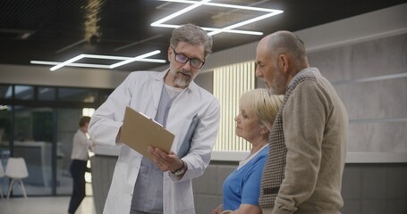 Obraz premium Elderly couple stands with doctor in clinic lobby near reception. Doctor speaks with patients, shows medical examination or test results. Medical staff and people in medical center. Healthcare system.