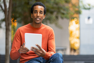 Fototapeta premium Portrait of smiling African American man holding digital tablet, reading e book, sitting on urban street, outdoors, copy space. Handsome businessman checking mail, text message. Technology concept