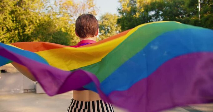 Portrait of a happy girl in a purple top and striped pants who rides on roller skates in a skate park and holds an LGBT flag that flutters in the wind