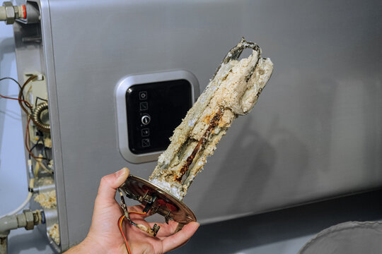 Heating element of an electric water heater with a rusty anode and a tube covered with scale. Serviceman holds a damaged part in his hand. Service and repair.