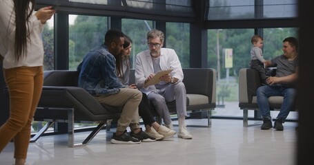 Diverse people sit on couches in clinic lobby area, wait for doctors appointment. Doctor talks to...