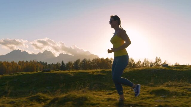 CLOSE UP, LENS FLARE: Female runner jogging in sunset light along a scenic trail. Last rays of golden autumn sun spill over beautiful alpine landscape as she runs along a picturesque meadow hill.