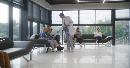 Diverse people sit on sofas in clinic lobby area, wait for appointment with doctor. Doctor speaks with elderly couple about medical test results. Waiting area in modern medical center. Healthcare.