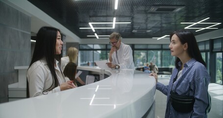 Asian female administrator talks with patient. Adult woman stands near reception desk in clinic lobby area, asks information, makes appointment with doctor. Medical staff work in modern hospital.