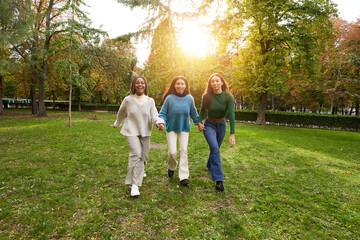 Naklejka na ściany i meble Three multiracial young girls stroll happily holding hand outdoors in the park. Happy and joyful women with friends on vacation on a sunny spring day. Beautiful colleagues enjoying the weekend.