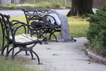 Anonymous homeless man and possible drug addicts sleeping on a New York City park bench with blanket over head - obrazy, fototapety, plakaty