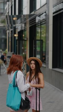 In the urban setting, two companions with suitcases and handbags discuss things with each other.