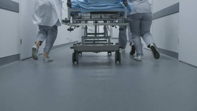 Doctors and nurses run and push stretcher with seriously injured patient to operating room. Medics save human life. Close up of medical personnel legs. Bright medical facility corridor. Slow motion.