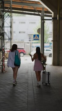 Two friends, both full of life, are seen walking with suitcases and handbags on the city streets.