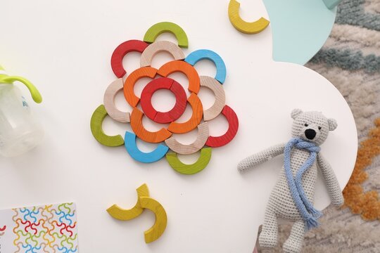Colorful wooden pieces of playing set and toy bear on white table, flat lay. Motor skills development