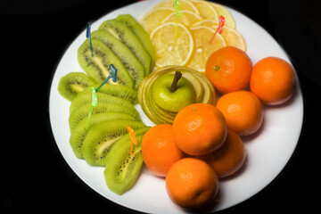 Fruit white plate with tangerines, kiwi, sliced apple and lemon slices on background isolated black
