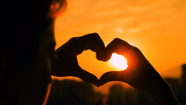 Close up view: silhouettes of woman hands are forming a heart shape sign against the sunset or sunrise orange sky - sun lens flares. Love, romantic, gesture and positive concept