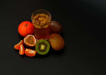 Tropical fruit juice with seeds on a black background, next to pieces of ripe passion fruit, tangerine and kiwi.