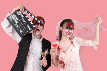 Young couple dressed for Halloween as zombies with movie clapper and cocktails on pink background
