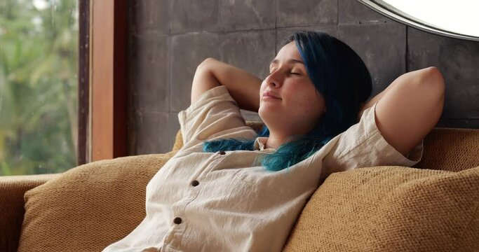 Young body positive woman with blue dyed hair sits half-lying on couch, leaning back on soft backrest and raising her hands behind her head. Stretching body and yawning demonstratively