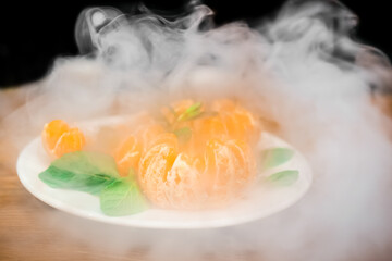 Peeled citrus tangerine with mint petals on a plate in white smoke from a hookah or vape of an...