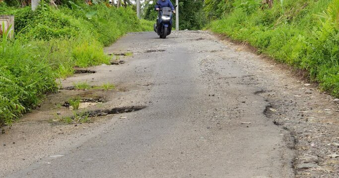 Motor scooter ride towards at small road with large potholes on left side. Driver man try to move around dangerous places. Bad condition of asphalt at local village road in countryside of central Bali