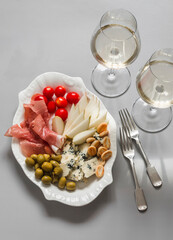 Tapas plate - ham, cheese, crackers, olives, cherry tomatoes and two glasses of white wine on a gray background, top view