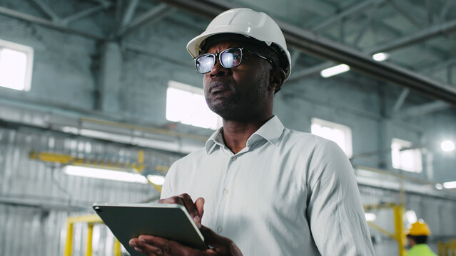 Careful concerned hard-working African-American male wearing white helmet marvelous glasses and white shirt looking around. Man using his superb tablet.