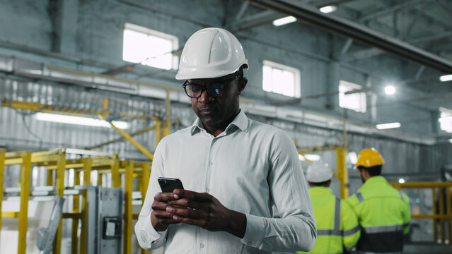 Interested responsible tall African-American male walking on industrial workplace looking in different directions. Man noting typing information using modern smartphone.