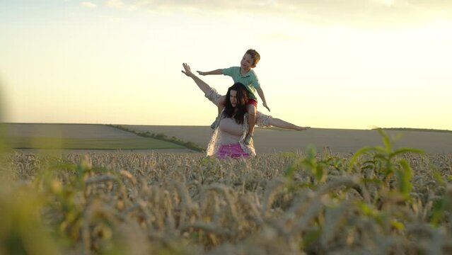 Mother and son fly with arms folded like an airplane over a wheat field. Happy childhood, parental love on farm agro fields in the village. High quality 4k footage