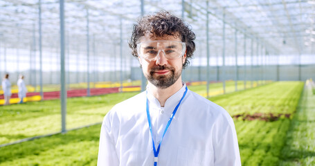 Portrait of smiling male scientist in lab coat and protective glasses looking at camera while standing at greenhouse farmland. Bearded man testing new environmental technology at hydroponic farm.