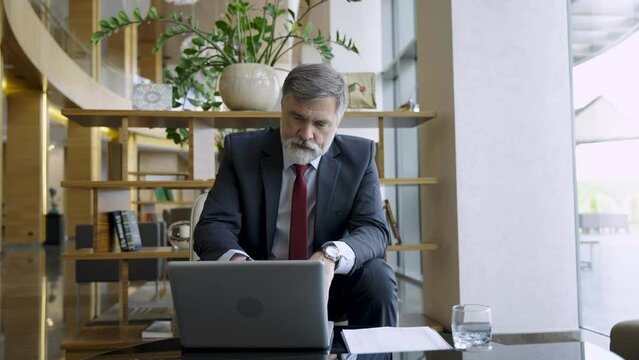 Businessman sitting at table in hotel lobby, restaurant or cafe, working on laptop computer. Mature age man in business casual, confident happy smiling