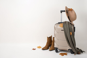 Female autumn adventure with luggage. Side view of a chic suitcase, comfy scarf, ankle boots, felt hat, pine cone, and maple leaves against a white background with space for text or ads