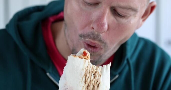 Hungry man eats shawarma sitting in light premise on blurred background. Lunch break and eating fast food at home concept slow motion