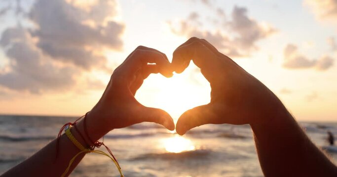 Couple makes heart sign against setting sun illuminating sea water. Tourists enjoy couple summer holiday at ocean resort slow motion