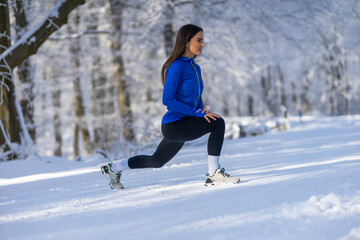 Fototapeta na wymiar Young sportswoman in perfect shape stretching leg in nature at snowy winter day.
