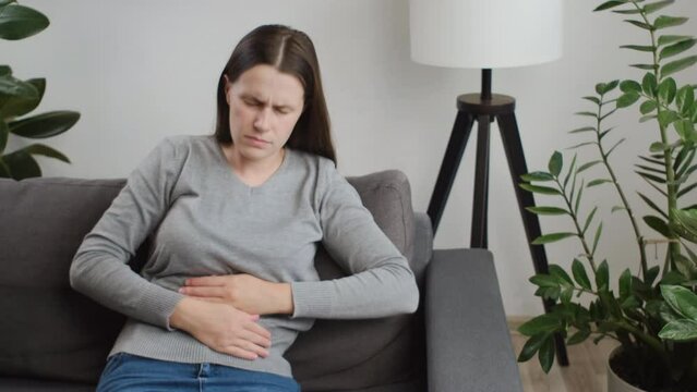 Upset female holding belly and feeling abdominal menstrual pain or bowel and digestion problems. Unhealthy sad young caucasian woman suffering from stomach ache sits on sofa. Food poisoning concept
