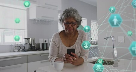 Animation of connected icons forming globes, senior african american woman using laptop in kitchen - Powered by Adobe