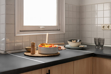 Salad with tomatoes stands on a round straw mat next to peppers and dishes on a black countertop in the kitchen on the background of white tiles in the hotel
