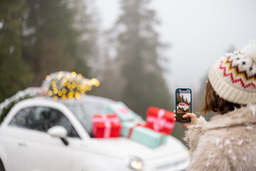 Woman makes a photo on phone of beautiful car decorated with presents and Christmas tree on rooftop, while traveling by vehicle in mountains on winter holidays