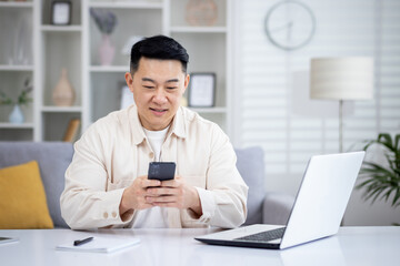 Mature Asian man sitting at table at home, man using phone, typing message on smartphone, app user and browsing internet in home cinema room, smiling contentedly.
