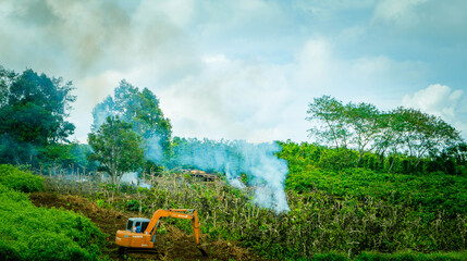 Yellow crawler excavator digs the ground against the blue sky with clouds, green trees and smoke. Construction work in the highlands.